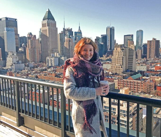 Eine Frau mit Kaffeebecher steht auf einer Dachterrasse vor der Skyline von Manhattan.