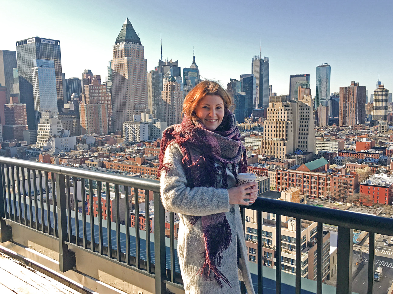 Eine Frau mit Kaffeebecher steht auf einer Dachterrasse vor der Skyline von Manhattan.