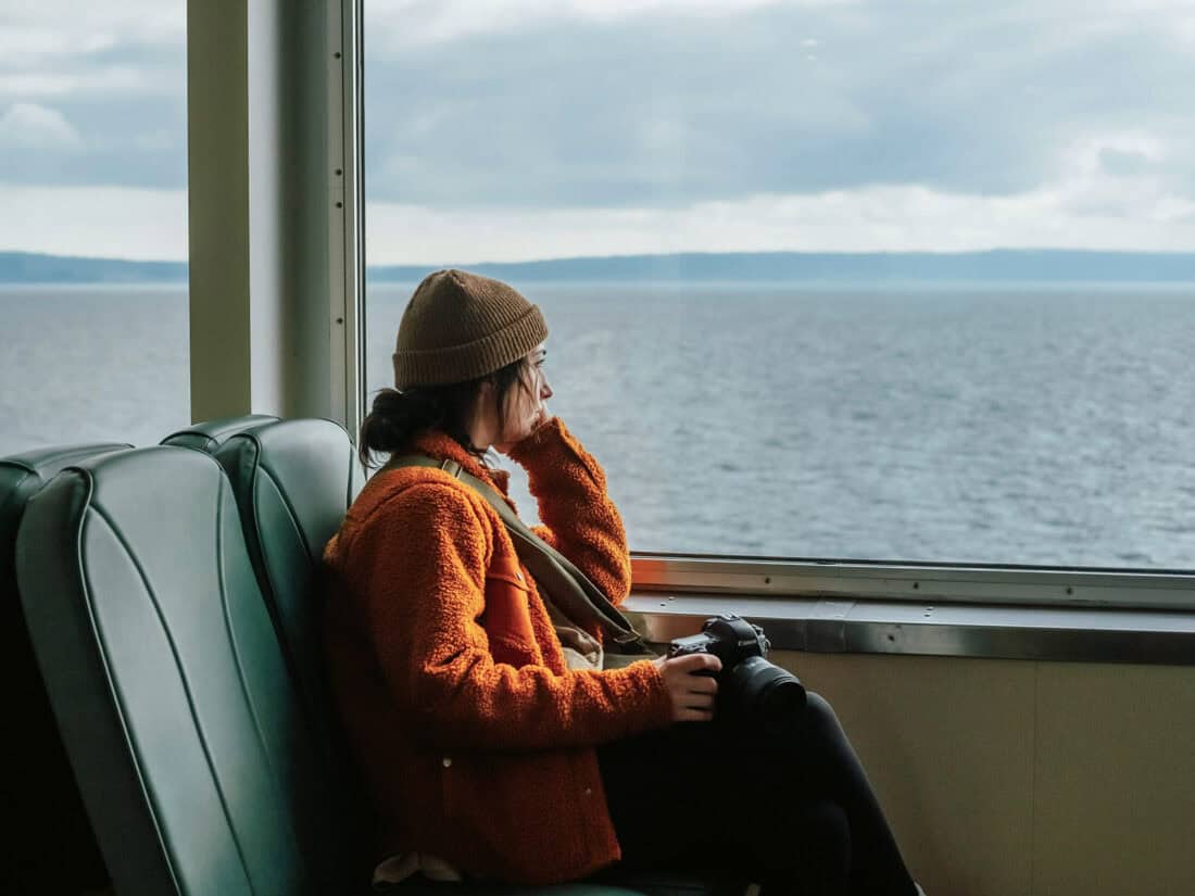 Alleinreisende Frau mit Kamera sitzt am Fenster in einem Boot und schaut nachdenklich aufs Meer.