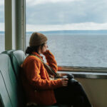 Alleinreisende Frau mit Kamera sitzt am Fenster in einem Boot und schaut nachdenklich aufs Meer.