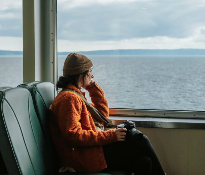 Alleinreisende Frau mit Kamera sitzt am Fenster in einem Boot und schaut nachdenklich aufs Meer.