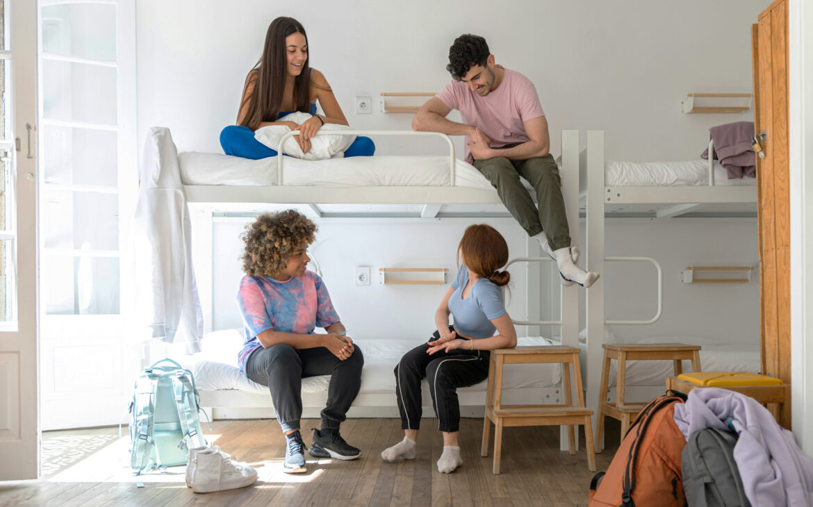Mehrbettzimmer in einem Hostel mit jungen Erwachsenen, die sich entspannt unterhalten und auf Etagenbetten sitzen.