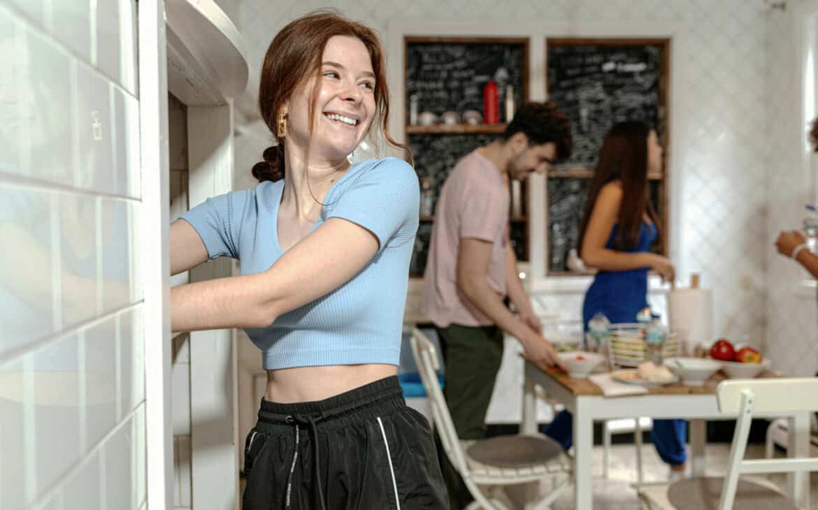 Alleinreisende Frau räumt in einer Hostelküche ein Regal ein, während andere Reisende im Hintergrund gemeinsam kochen und sich unterhalten.