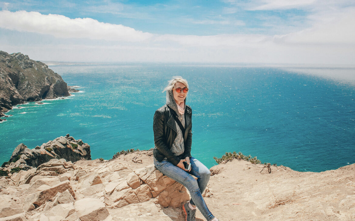 Frau über 50 mit Sonnenbrille und Lederjacke sitzt auf einem Felsen an einer Steilküste über dem türkisblauen Meer – Roadtrip allein
