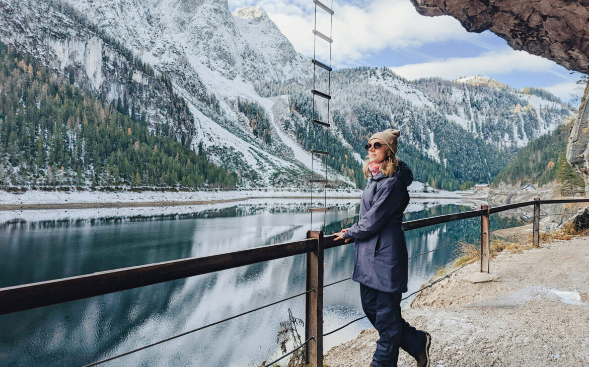 Frau über 50 in Wintermantel und Mütze steht an einem Geländer vor einem Bergsee mit verschneiten Gipfeln – Wanderreise allein